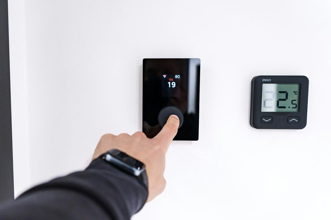 A hand adjusts a digital thermostat on a white wall.