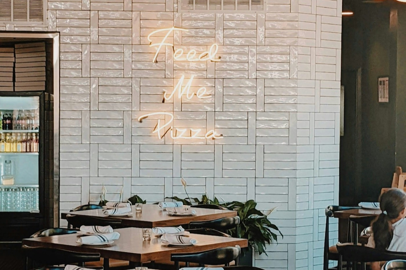 A restaurant dining room with set tables, plants and a neon sign on a white tiled wall.