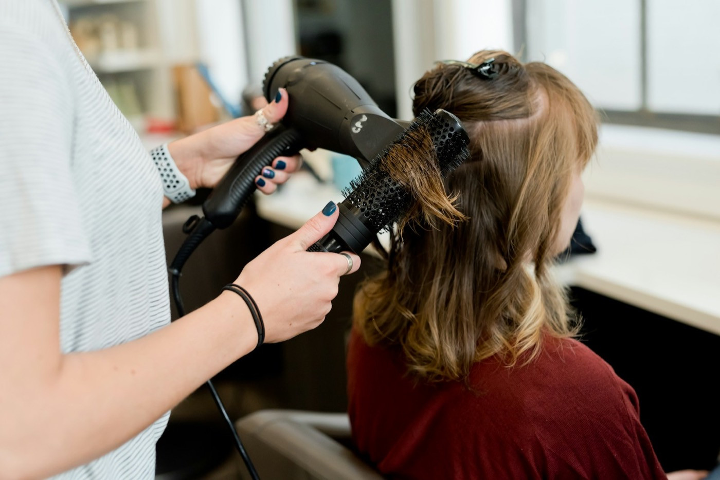 A hairstylist blow-dries a client’s shoulder-length hair with a round brush in a salon.
